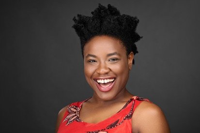 A joyful woman with a radiant smile poses in a vibrant red top against a dark background, exuding positivity and confidence in her headshot captured by the photographer.