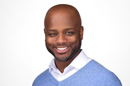 A confident man with a bald head and a friendly smile, captured by a headshot photographer, wearing a blue sweater over a collared shirt, against a neutral background.