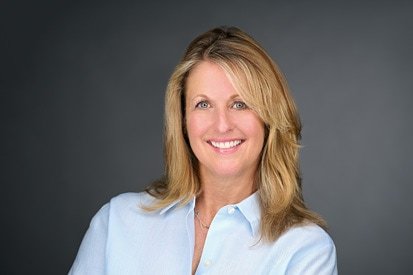 A confident professional woman with blonde hair smiling in a formal blue shirt against a gray background, projecting a friendly and approachable demeanor, captured by a talented headshot photographer.
