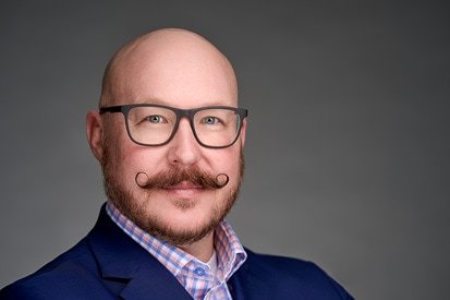 A confident man with a styled handlebar mustache, wearing glasses and a smart blue blazer over a checkered shirt, poses against a neutral gray background for his headshot photographer.