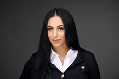 A professional woman with long black hair, wearing a smart black blazer adorned with buttons, poses against a grey backdrop for her headshot photographer, exuding confidence and poise.