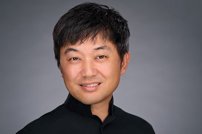 A smiling Asian man with short black hair wearing a black shirt, against a smooth grey background, portraying confidence and approachability in his professional headshot.