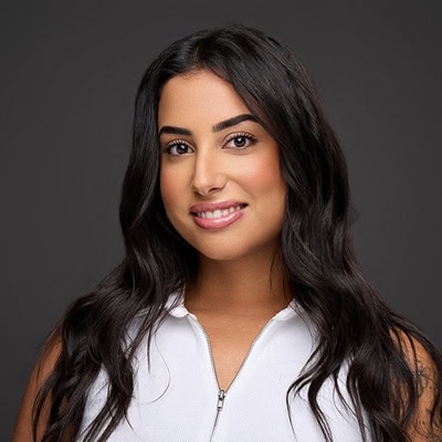 Portrait of a smiling woman with long dark hair, captured by our team of headshot photographers. She is wearing a white zip-up top against a gray background. Her makeup is natural, highlighting her gentle smile.