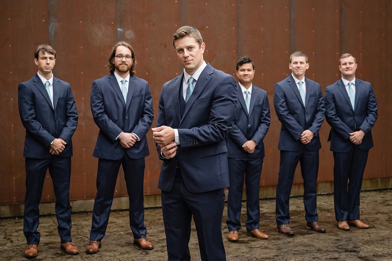 A group of seven men in matching dark blue suits and brown shoes, demonstrating how to button a suit with a confident stance against a rustic brown background. The man in the foreground is adjusting his cufflinks