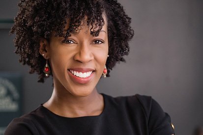 A confident woman with a radiant smile, sporting curly short hair, wearing a black top and red earrings, stands before a blurred background suggesting a professional setting by a professional headshot photographer.