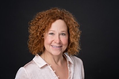 A radiant woman with curly auburn hair smiles gently at the camera, her joy subtly reflected in her sparkling eyes, set against a muted background. Captured by a professional headshot photographer.