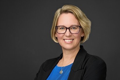 A professional woman with blonde hair and glasses, smiling confidently in a black blazer and blue top, against a grey background. Captured by a skilled headshot photographer, she conveys an approachable