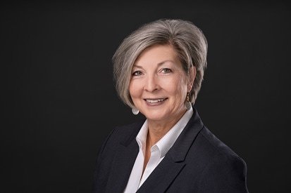 A professional headshot of a smiling, senior businesswoman with silver hair, dressed in a smart black blazer and white shirt, set against a dark gray background.